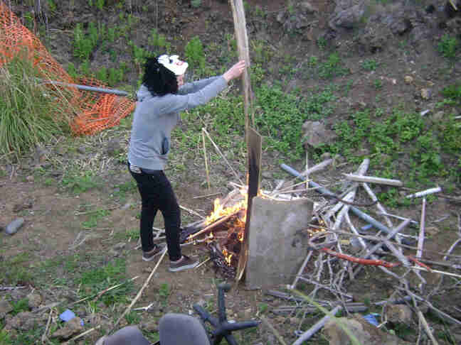 Right side view of a person, wearing a costume mask over their head, setting wood on a burning campfire in a pit